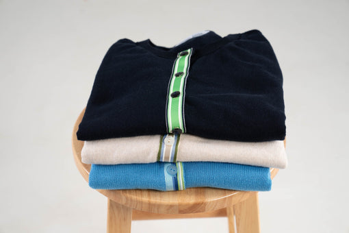 Noyac Knits premium cotton crew neck cardigans with ribbon trim in navy, tan and French blue shown folded on a stool on white background. These cardigans can be worn in early fall and as summer to fall transition pieces.