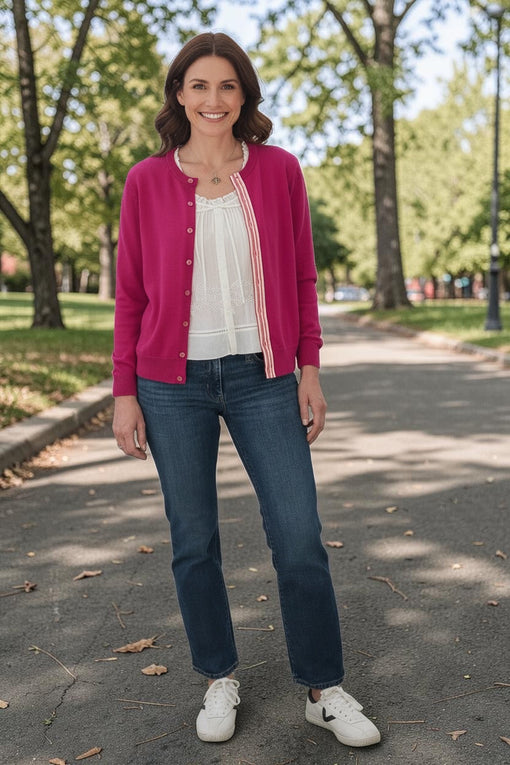 Woman wearing a raspberry pink Noyac Knits Grace classic cardigan with striped grosgrain ribbon trim over a white blouse, styled with dark wash jeans and white sneakers, standing on a tree-lined park path in fall.