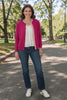 Woman wearing a raspberry pink Noyac Knits Grace classic cardigan with striped grosgrain ribbon trim over a white blouse, styled with dark wash jeans and white sneakers, standing on a tree-lined park path in fall.