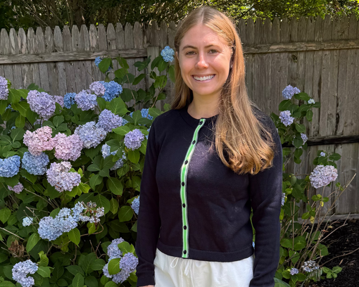 Woman wearing Noyac Knits Julie Crop Cardigan in Navy Cotton and trimmed with vintage-inspired grosgrain ribbon in stripes of navy, white and kelly green.  Styled over white linen pants.  Hydrangeas and wooden fence in background.