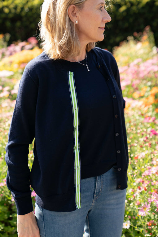 Woman in garden setting wearing Julie classic cardigan in navy merino with ribbon trim in stripes of navy, white and navy over matching sleeveless shell in navy merino.