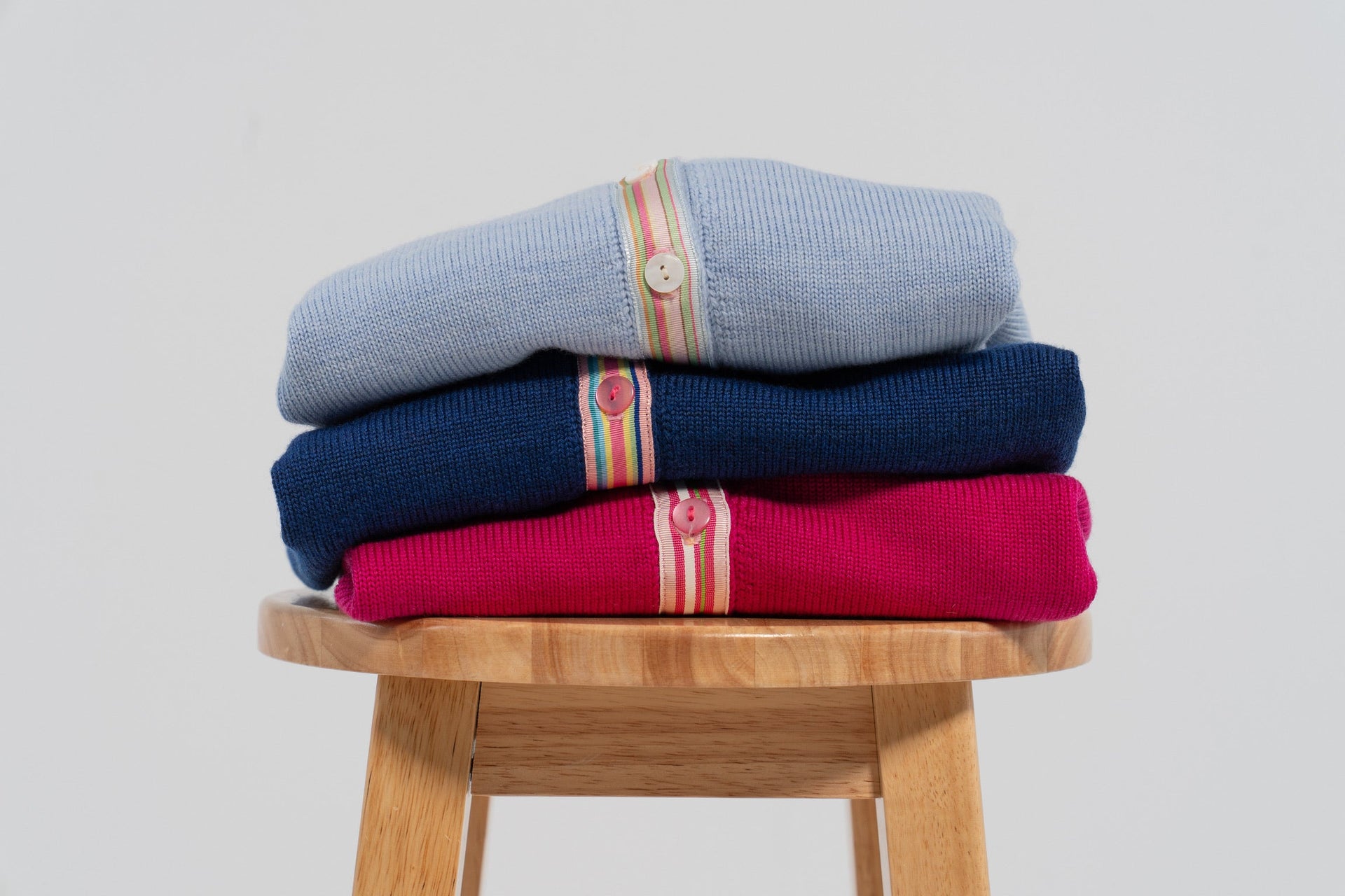 Stack of three cardigans in light blue, cobalt blue and raspberry pink and plain background.
