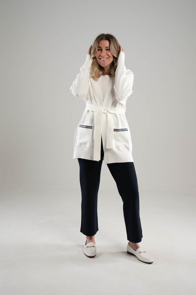 woman wearing long white cardigan with navy and white striped ribbon trimmed pockets and navy ankle length knit pants on plain background