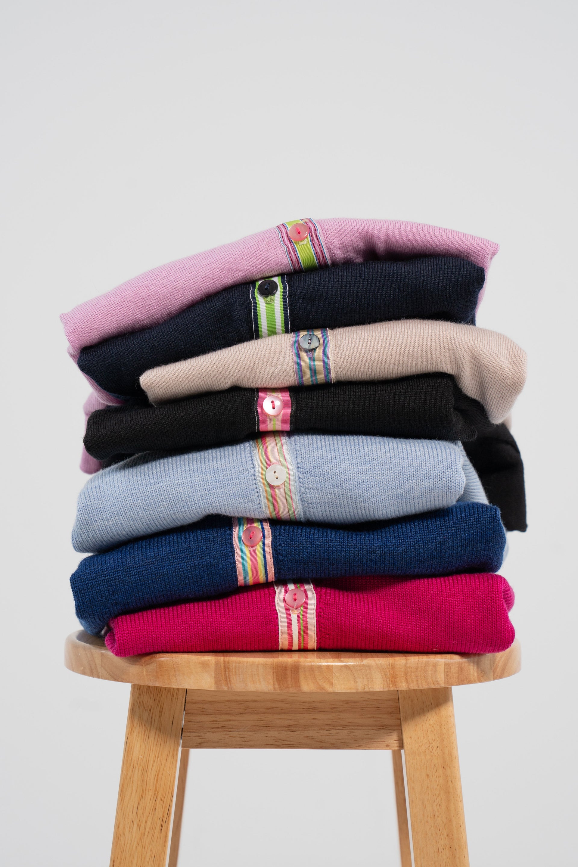 Stack of colorful cardigans on wood stool and plain background.