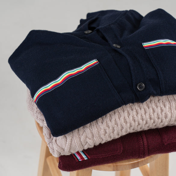 Three sweaters stacked on stool with plain background.  Sweater on top is navy with striped ribbon trim on pockets.  Middle sweater is beige cableknit.  Bottom sweater is burgundy with striped ribbon trim pocket.