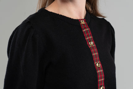 close up of black sweater with red, black and gold tartan plaid ribbon trim and gold buttons on plain background.