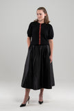Woman wearing black puff-sleeve cardigan with red tartan plaid ribbon trim and gold buttons, black skirt and black patent sling-back shoes on plain background.
