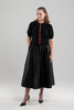 Woman wearing black puff-sleeve cardigan with red tartan plaid ribbon trim and gold buttons, black skirt and black patent sling-back shoes on plain background.