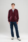 Man wearing Noyac Knits bomber-style sweater in burgundy color with signature striped ribbon-trimmed pockets -- knit in premium, ultrasoft merino wool -- styoed with  light blue t-shirt, jeans and sneakers on plain background.