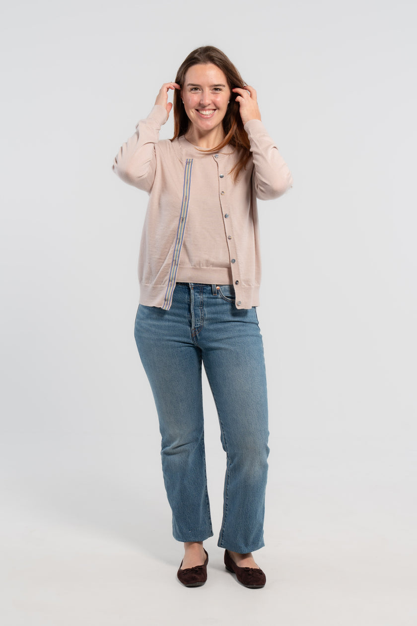 Woman wearing Noyac Knits Isabelle Classic Cardigan in light beige trimmed with signature grosgrain ribbon in stripes of tan, purple, lavender, and teal and Japanese mussel shell buttons -- knit in lightweight premium merino -- styled with matching merino wool crew neck sleeveless shell, jeans, balle