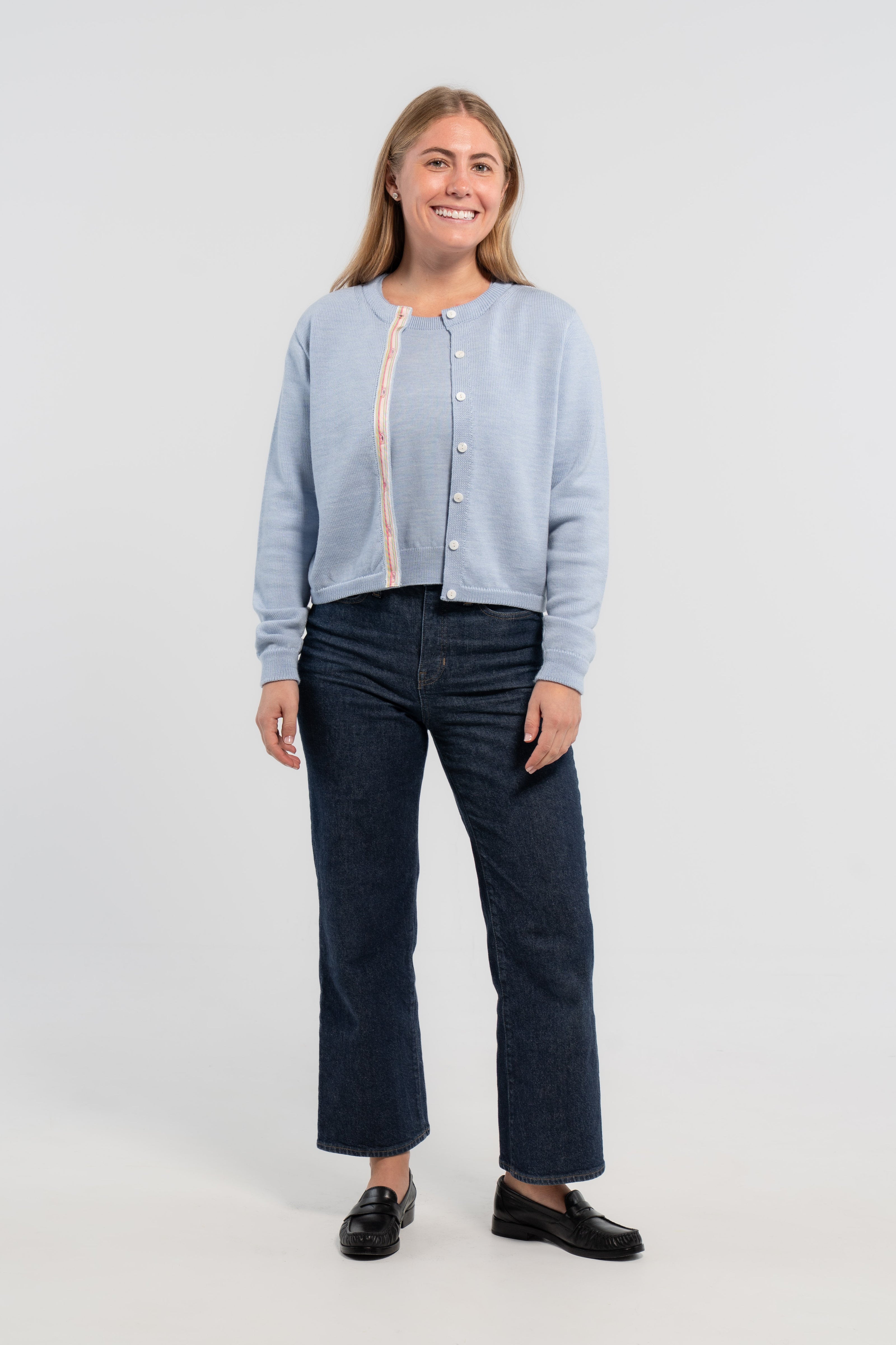 Woman wearing Noyac Knits Mary crop cardigan in soft, premium merino and trimmed with white mother of pearl buttons and signature ribbon in stripes of pastel pink, blue and green striped grosgrain ribbon styled with jeans and loafers on plain background.