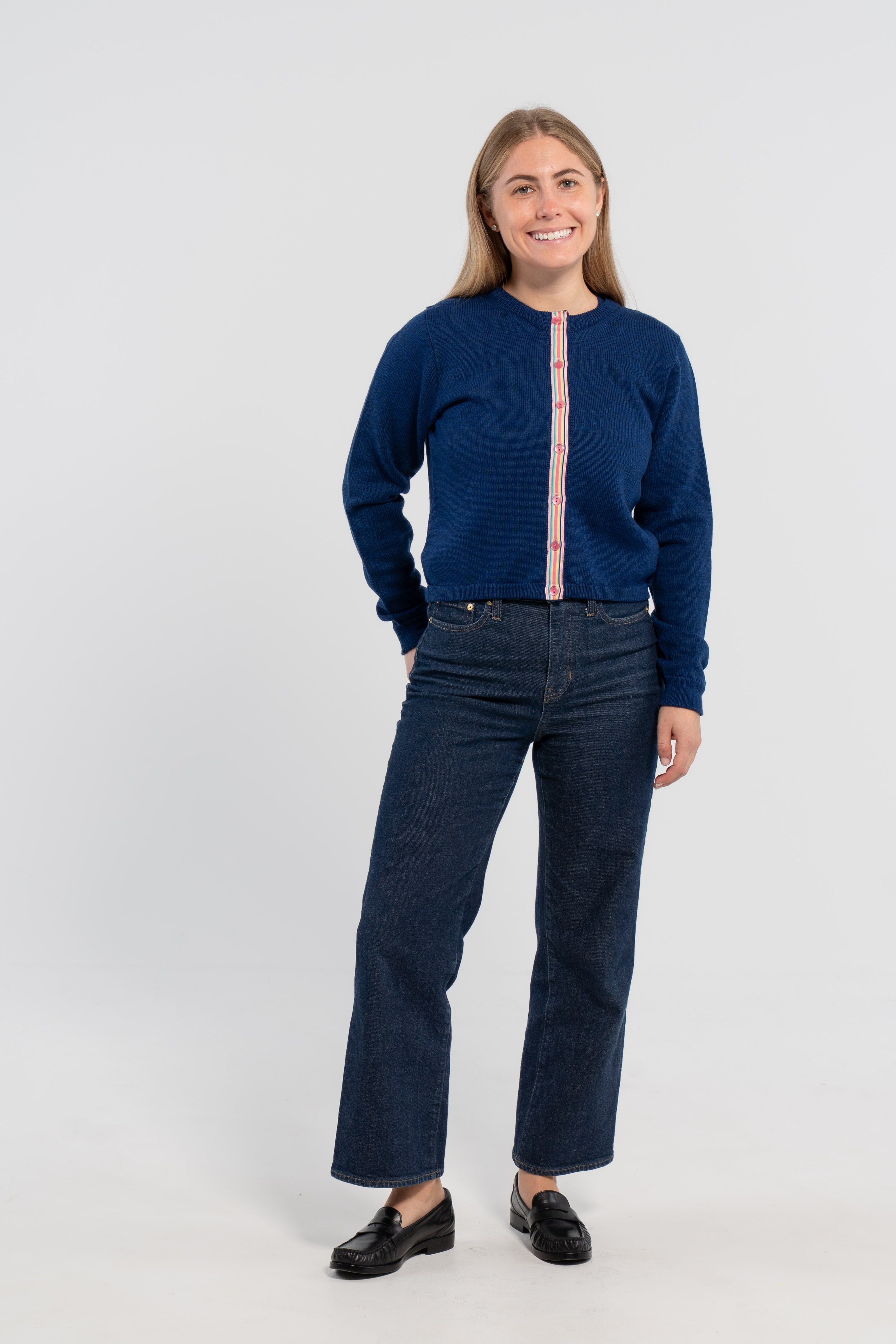 Woman wearing Noyac Knits Sissy Crop Cardigan in gorgeous cobalt blue trimmed with signature pink buttons and rainbow striped grosgrain ribbon -- knit in premium merino - styled with matching merino cobalt blue crew neck sweater shell, jeans, loafers on plain background.