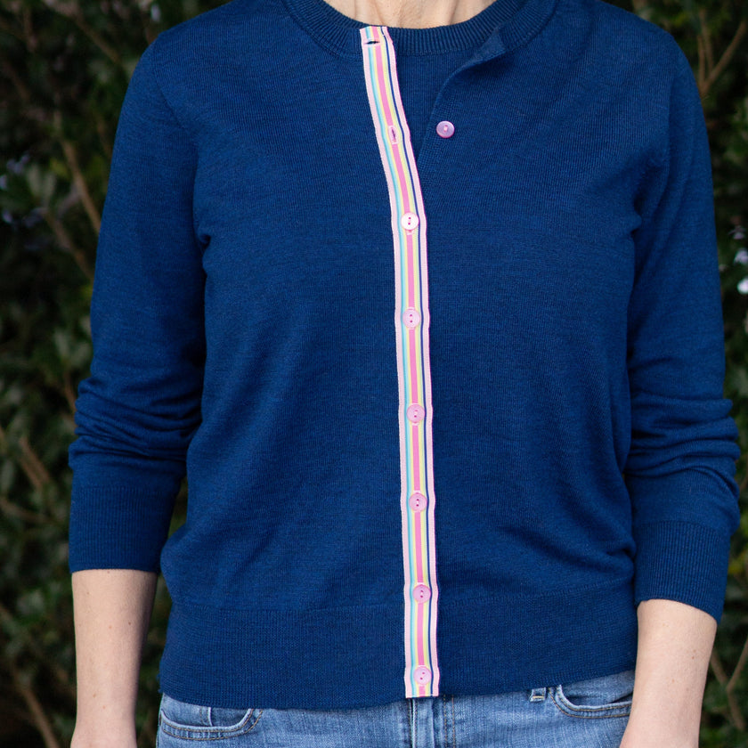 Woman wearing a Noyac Knits Sissy classic cardigan in gorgeous cobalt blue with signature pink rainbow striped ribbon trim  and pink buttons made in preimium lightweight merino styled with jeans against a green leafy background