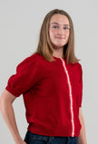 Woman wearing red Noyac Knits Rhoda short, puff-sleeve cardigan with signature ribbon trim in red, pink and white stripes   and white mother of pearl buttons, knit is soft, lightweight merino and styled with jeans on plain background.