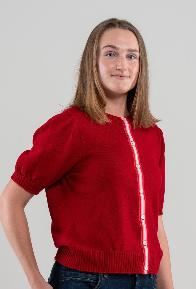 Woman wearing red Noyac Knits Rhoda short, puff-sleeve cardigan with signature ribbon trim in red, pink and white stripes   and white mother of pearl buttons, knit is soft, lightweight merino and styled with jeans on plain background.