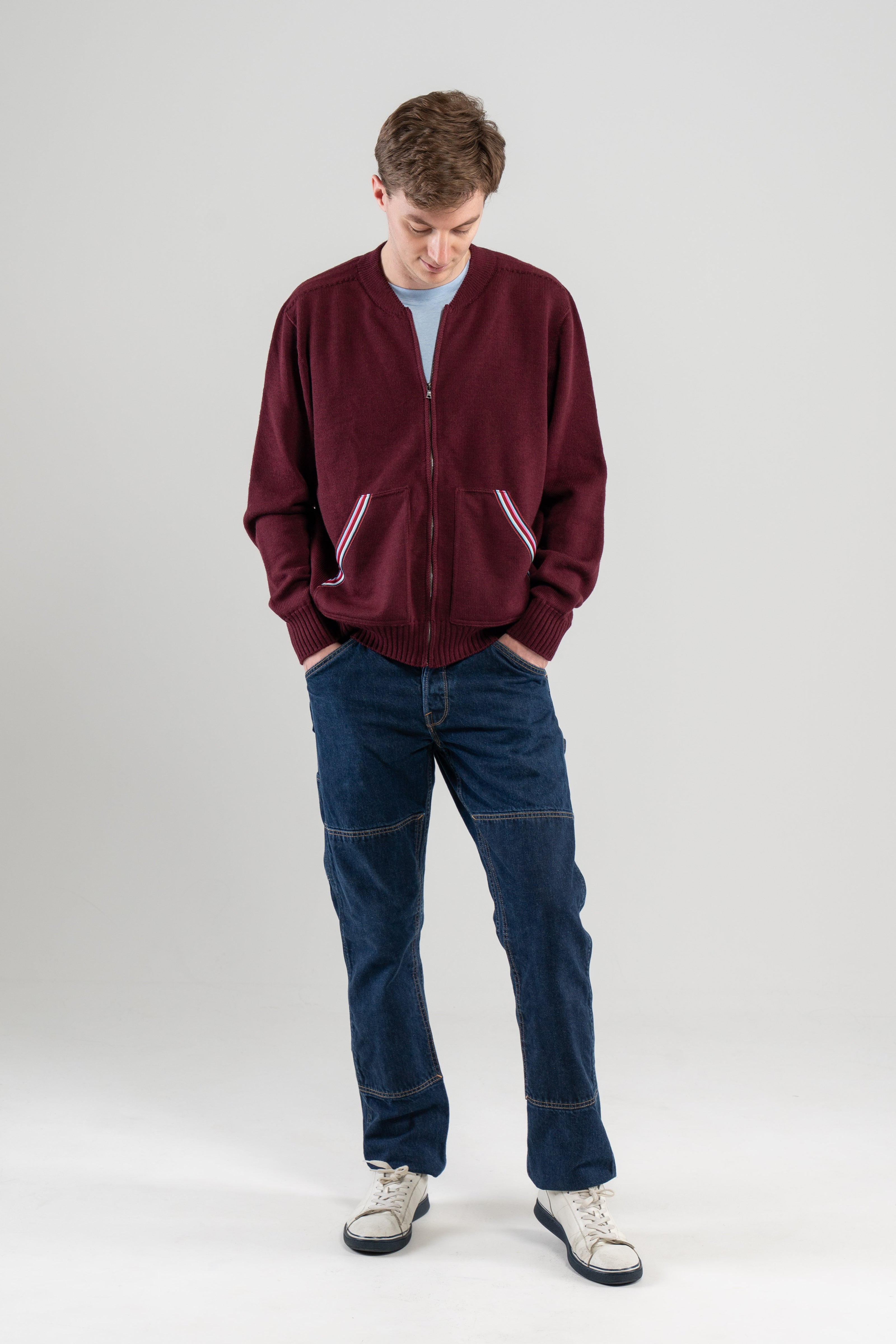 Man wearing Noyac Knits bomber-style sweater in burgundy color with signature striped ribbon-trimmed pockets -- knit in premium, ultrasoft merino wool -- styoed with  light blue t-shirt, jeans and sneakers on plain background.