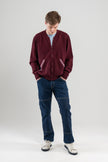 Man wearing Noyac Knits bomber-style sweater in burgundy color with signature striped ribbon-trimmed pockets -- knit in premium, ultrasoft merino wool -- styoed with  light blue t-shirt, jeans and sneakers on plain background.