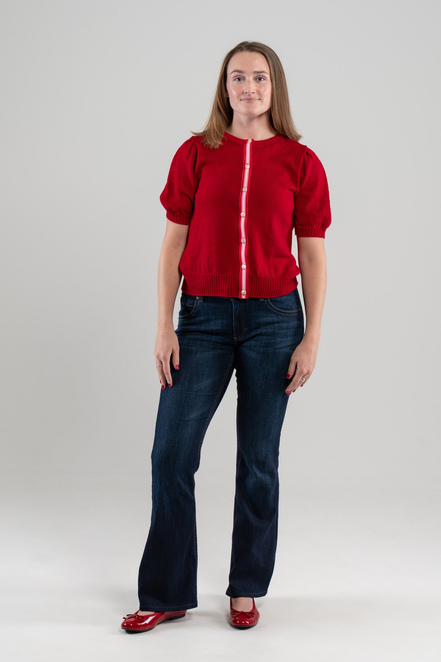 Woman wearing red puff-sleeve cardigan with red, pink and white striped ribbon trim and white buttons, jeans, red shoes on plain background.