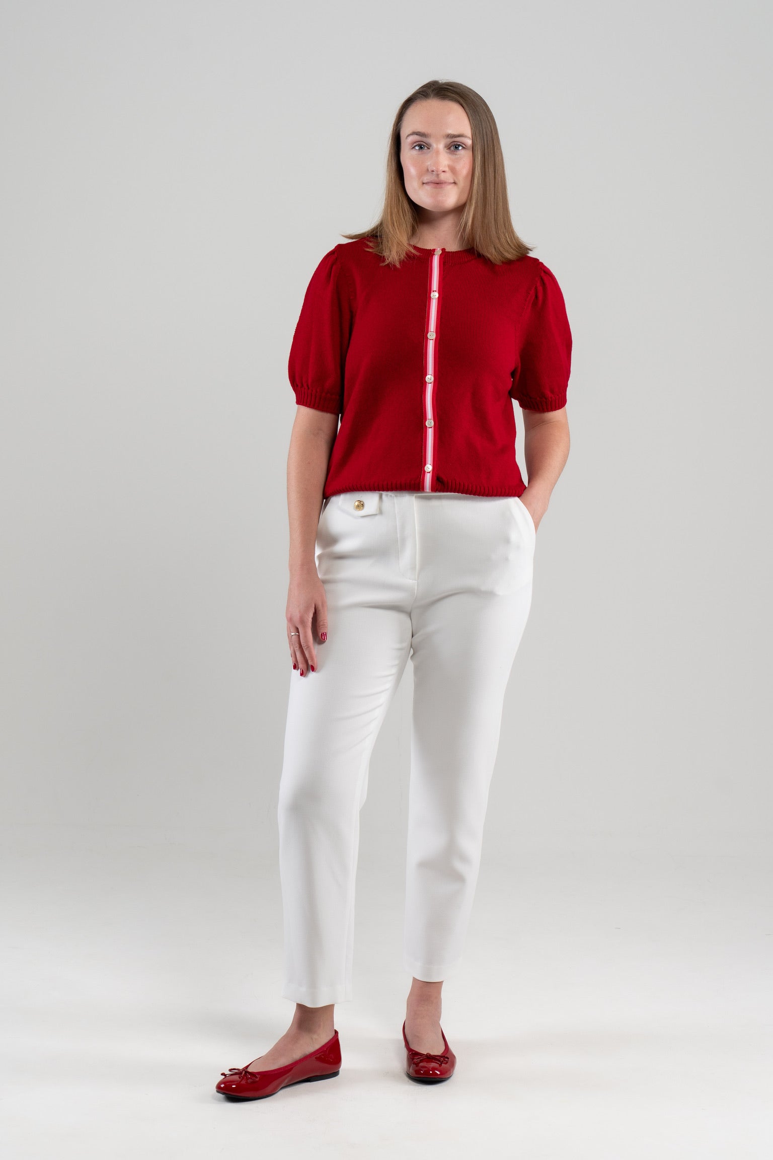 Woman wearing red puff-sleeve cardigan with red, pink and white striped ribbon trim and white buttons, white pants, red shoes on plain background.