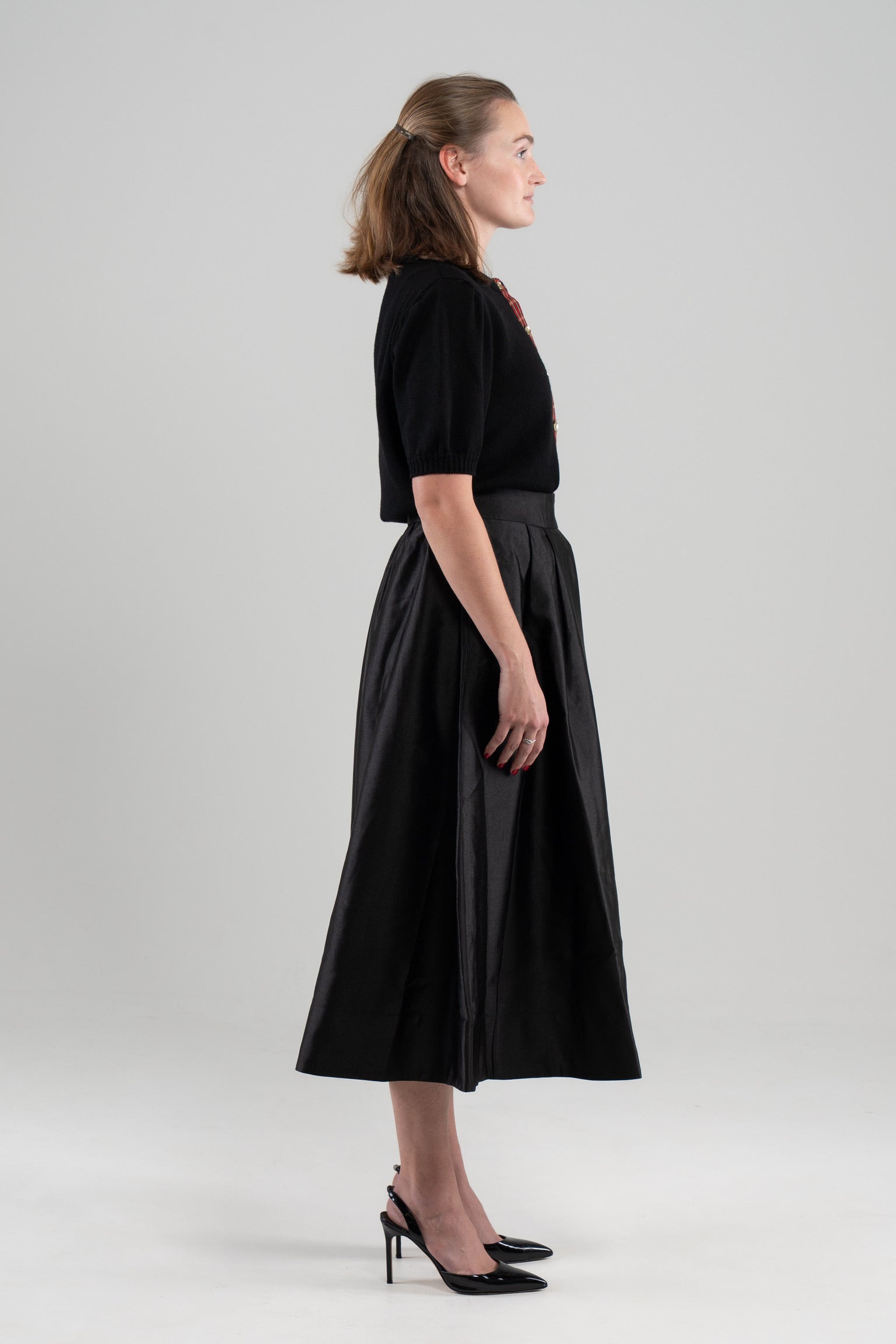Side view of woman wearing black puff-sleeve sweater and black skirt and black patent sling-back shoes on plain backbround.
