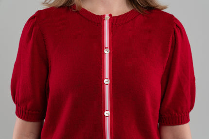Close up of red puff-sleeve cardigan with red, pink, and white striped grosgrain ribbon trim and white buttons on plain background.