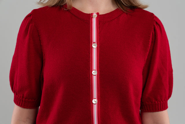 Close up of red puff-sleeve cardigan with red, pink, and white striped grosgrain ribbon trim and white buttons on plain background.