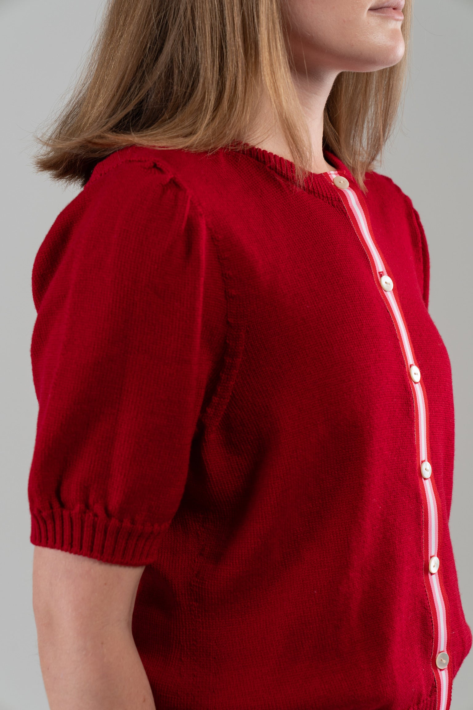 Side view close up of woman wearing red puff-sleeve cardigan with red, pink, white striped ribbon trim and white buttons on plain background.