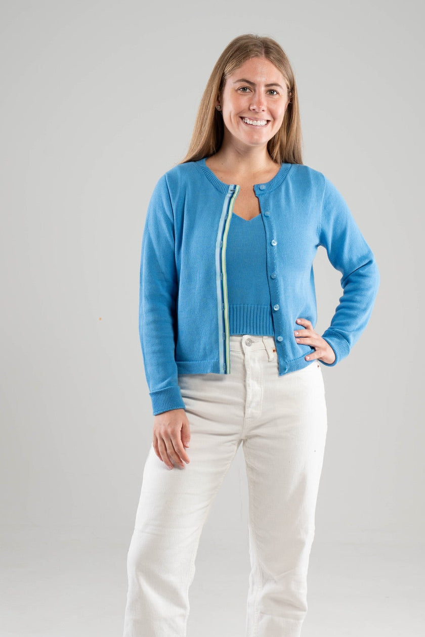 Woman wearing a french blue cardigan  with ribbon trim in green and blue stripes and white pants on a gray background