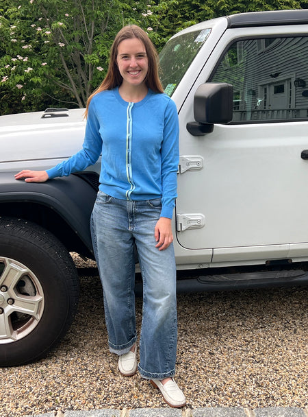 Woman in Noyac Knits sky blue Allison ribbon cardigan made in lightweight, breathable Egyptian cotton, styled with barrel jeans and white loafers standing next to a white Jeep on a gravel driveway.