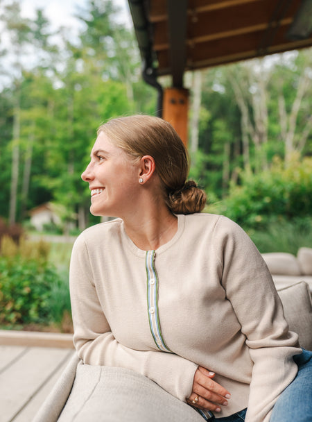 Woman sitting on outdoor couch in tan cardigan with striped ribbon trim with blurred green  trees and buses in background