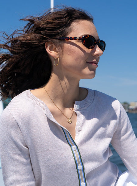 Woman on a boat wearing sunglasses with a clear blue sky and water background wearing Noyac Knits Lynn crop cardigan premium lightweight cotton in light tan with signature ribbon trim with blue, green, white and tan stripes and mother of pearl buttons.