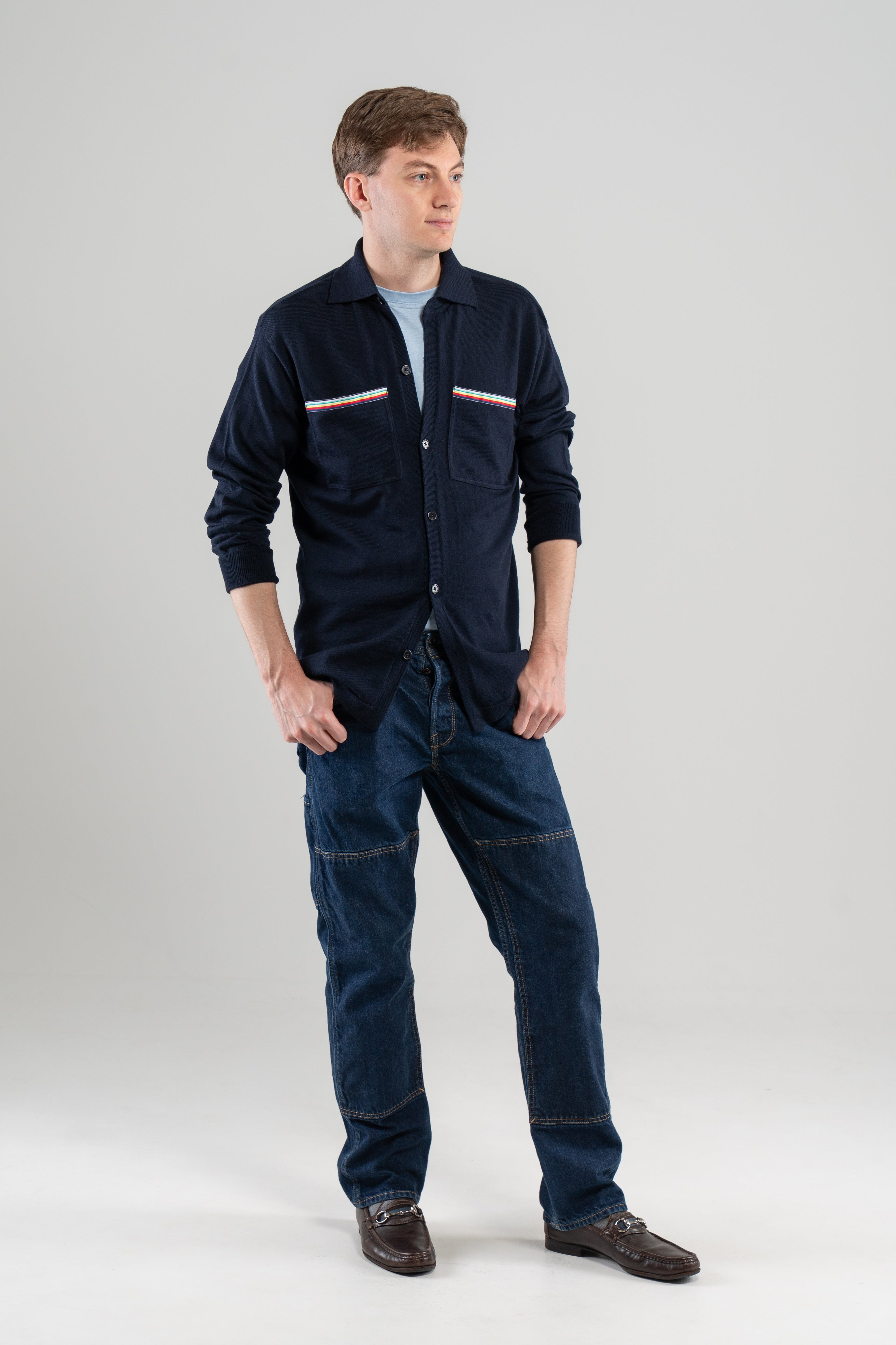 Man wearing Noyac Knits Finn knit overshirt in navy premium soft lightweight merino wool with signature ribbon detailing on pockets styled  over light blue t-shirt with jeans and oafers on plain background.