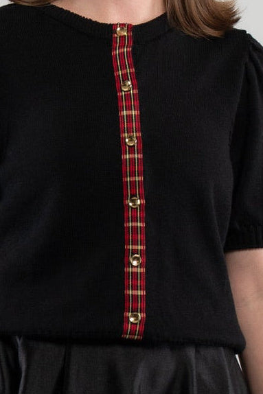 Close up of woman wearing black puff-sleeve cardigan with red tartan plaid ribbon trim and gold buttons on plain background.
