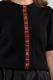 Close up of woman wearing black puff-sleeve cardigan with red tartan plaid ribbon trim and gold buttons on plain background.