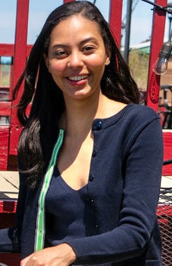 Woman wearing Noyac Knits navy blue crop cardigan in cotton with matching navy cotton v-neck shell sitting in front of a red fence outside.