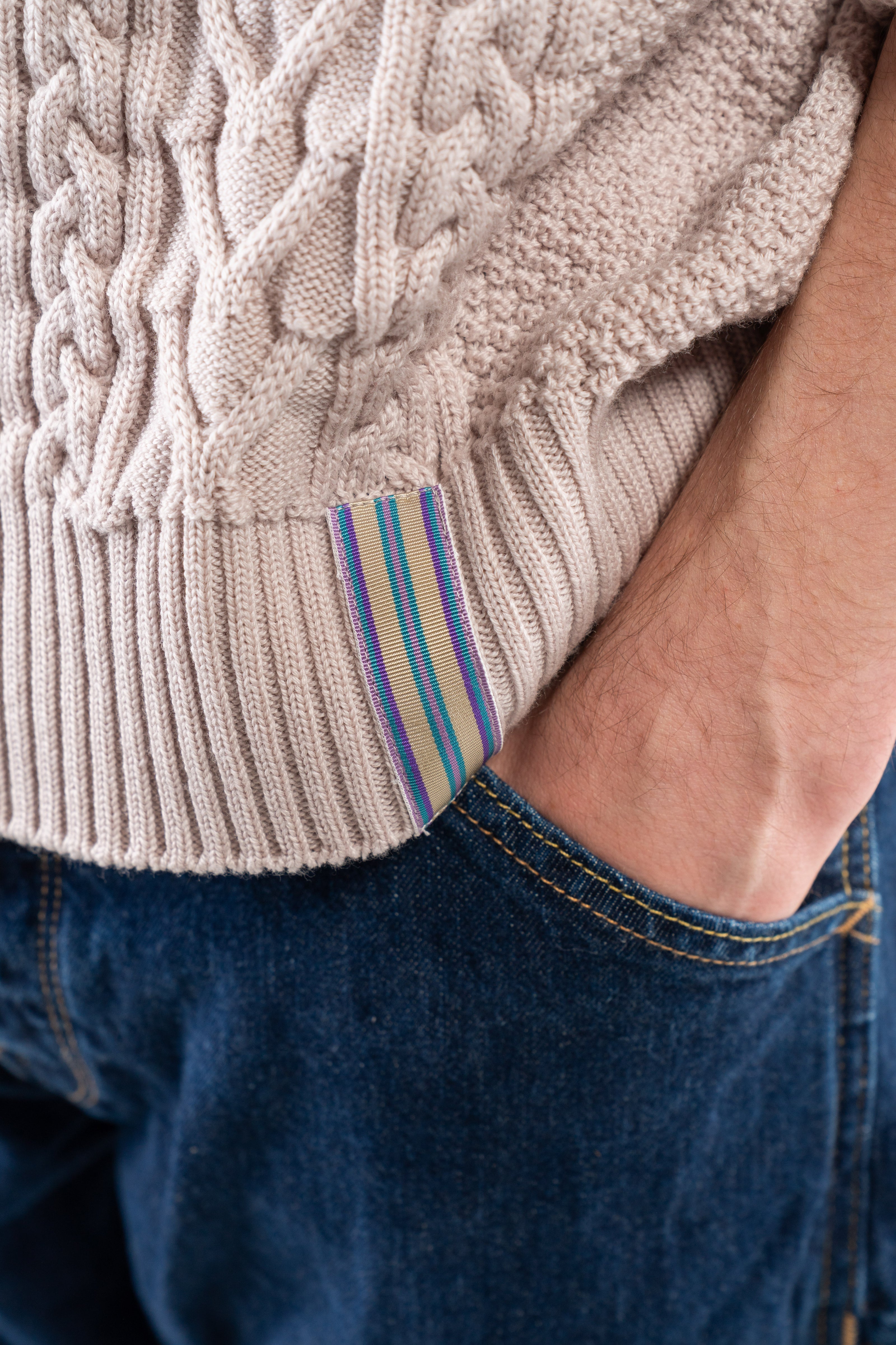 Close-up of Noyac Knits Cooper cableknit sweater in  beige premium merino wool showing ribbon detail on hem, with purple, tan, and teal stripes on ribbon 