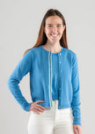 Woman wearing a Noyac Knits sky blue crop cotton cardigan trimmed in blue, green, and white striped ribbon with blue buttons styled over matching crew neck sleeveless shell with white jeans on plain background.