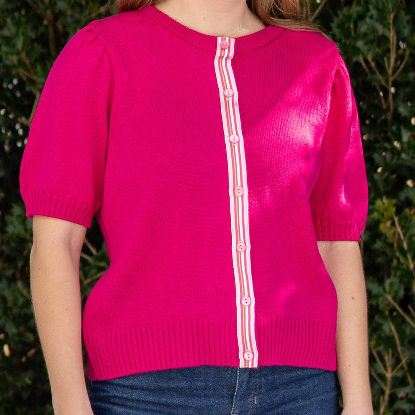 Woman wearing a dark pink Noyac Knits short puff sleeve ribbon cardigan trimmed with with pink, white, green striped ribbon and knit is premium, lightweight merino styled with blue jeans in outdoor setting with greenery in the background
