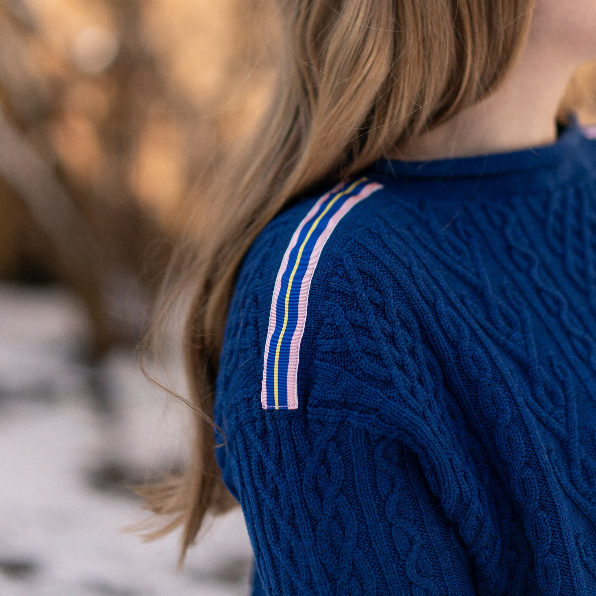 Close up of Noyac Knits cobalt blue boatneck pullover sweater with gorgeous cable detailing and signature ribbon trim in stripes of blue, pink and yellow on shoulder, knit in premium, soft merino wool shown on woman in natural setting and blurred background.