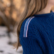 Close up of Noyac Knits cobalt blue boatneck pullover sweater with gorgeous cable detailing and signature ribbon trim in stripes of blue, pink and yellow on shoulder, knit in premium, soft merino wool shown on woman in natural setting and blurred background.