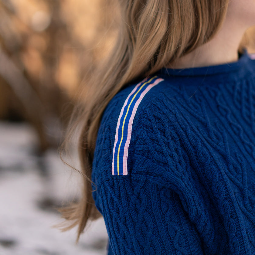 Close up of Noyac Knits cobalt blue boatneck pullover sweater with gorgeous cable detailing and signature ribbon trim in stripes of blue, pink and yellow on shoulder, knit in premium, soft merino wool shown on woman in natural setting and blurred background.