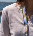 Close-up of a woman  wearing Noyac Knits light tan crop cardigan in lightweight cotton trimmed with green, blue, white and tan striped ribbon and mother of pearl buttons in outdoor setting.