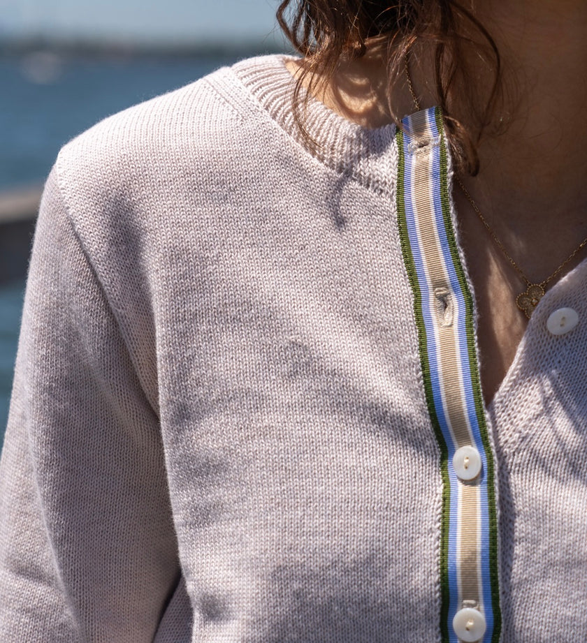 Close-up of a woman  wearing Noyac Knits light tan crop cardigan in lightweight cotton trimmed with green, blue, white and tan striped ribbon and mother of pearl buttons in outdoor setting.