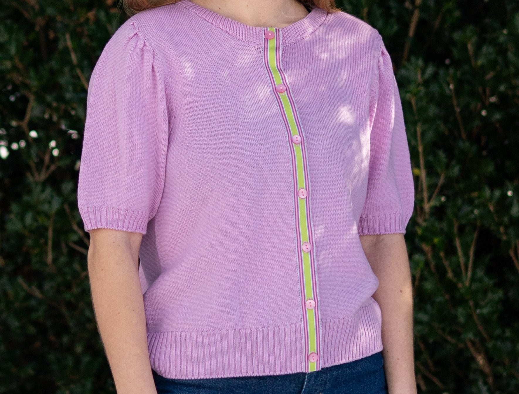 Woman wearing Noyac Knits lilac pink short puff-sleeve cardigan with pink and green ribbon trim, in premium, lightweight merino, styled with jeans against green leafy background