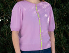 Woman wearing Noyac Knits lilac pink short puff-sleeve cardigan with pink and green ribbon trim, in premium, lightweight merino, styled with jeans against green leafy background