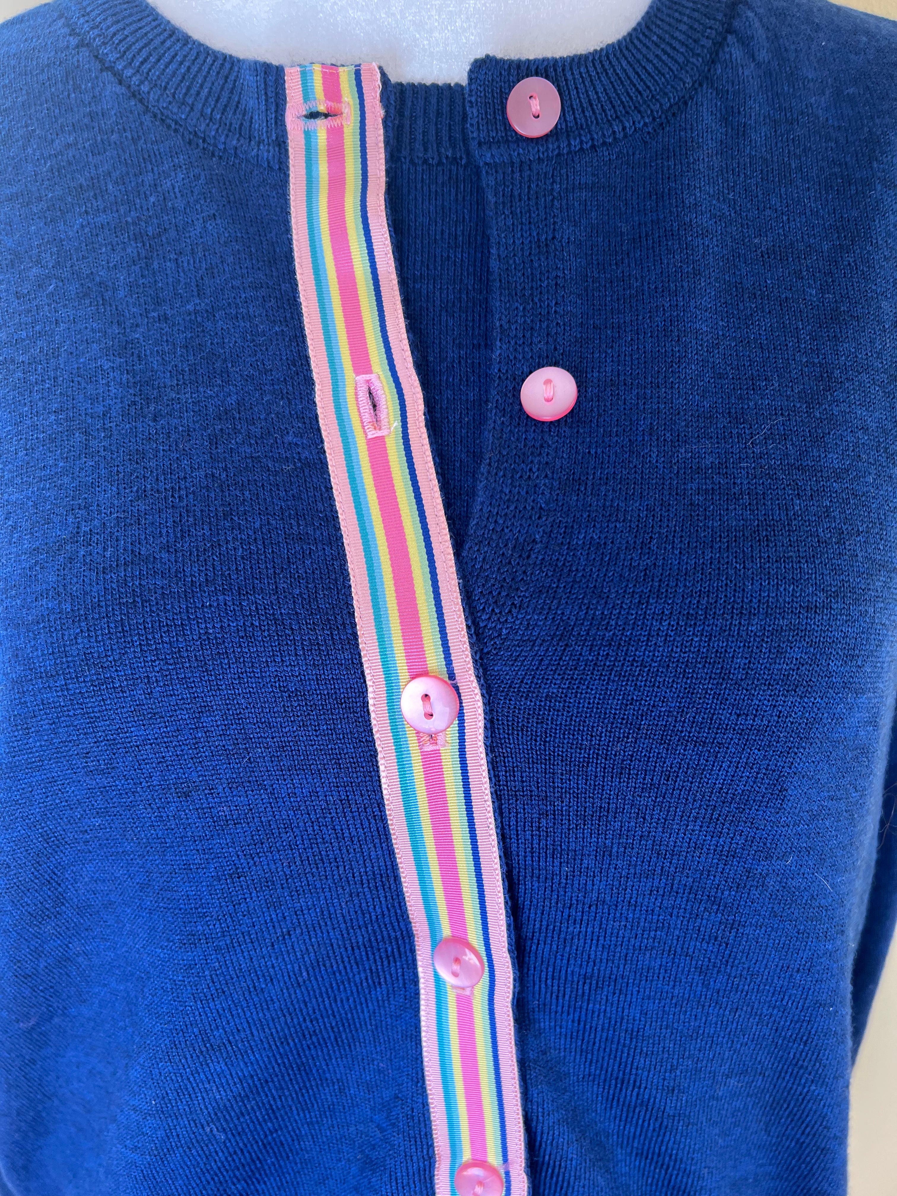 Close up of Noyac Knits Sissy classic cardigan in cobalt blue and trimmed with pink buttons and a rainbow-striped ribbon knit in lightweight premium merino