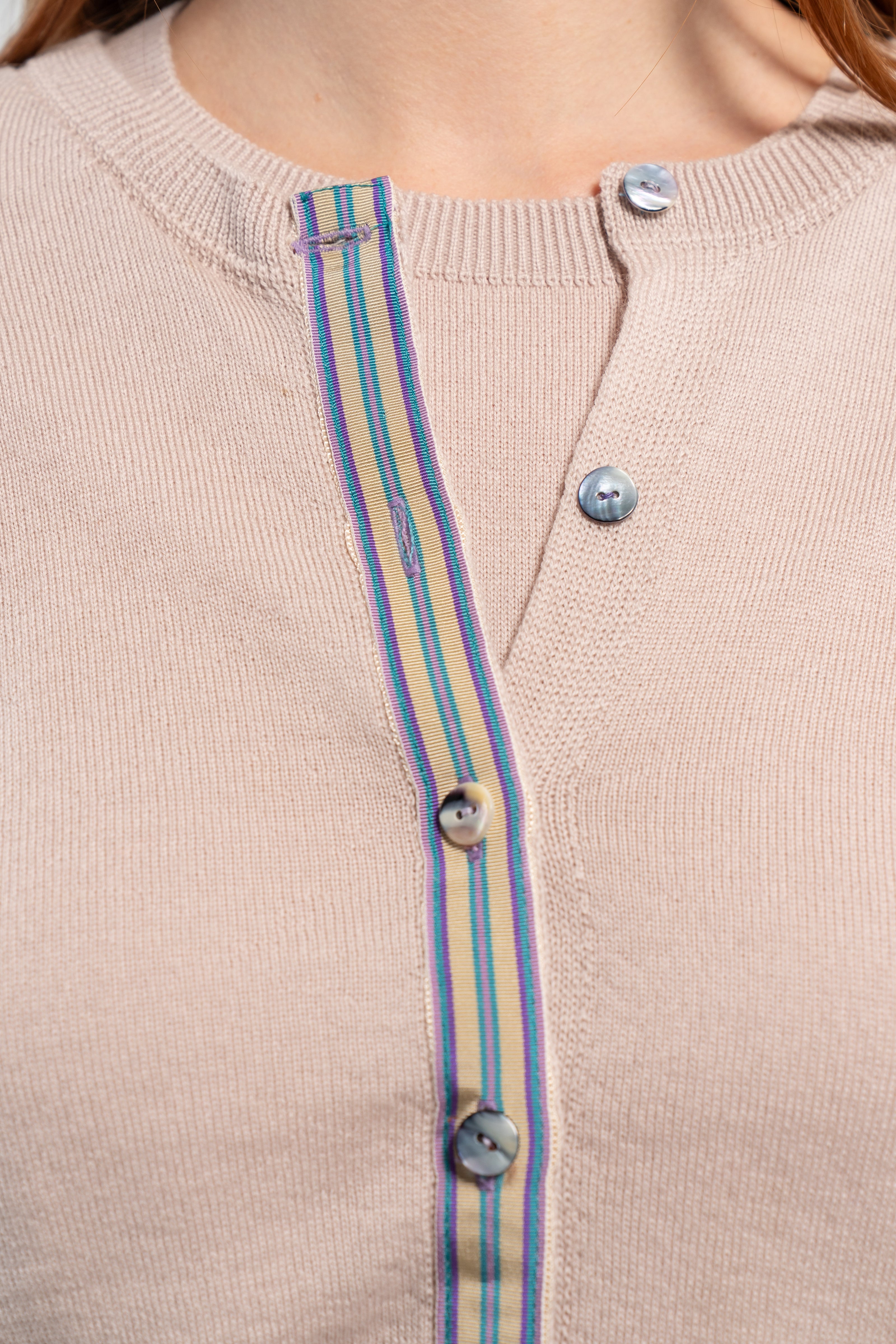 Close up of Noyac Knits Isabelle Classic Cardigan in light beige trimmed with signature grosgrain ribbon in stripes of tan, purple, lavender, and teal and Japanese mussel shell buttons -- knit in lightweight premium merino -- on plain background