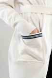 Close up Noyac Knits long belted white cardigan with a navy and white striped ribbon trimmed pocket on a plain background.  This cardigan is made from environmentally sustainable ecoviscose.