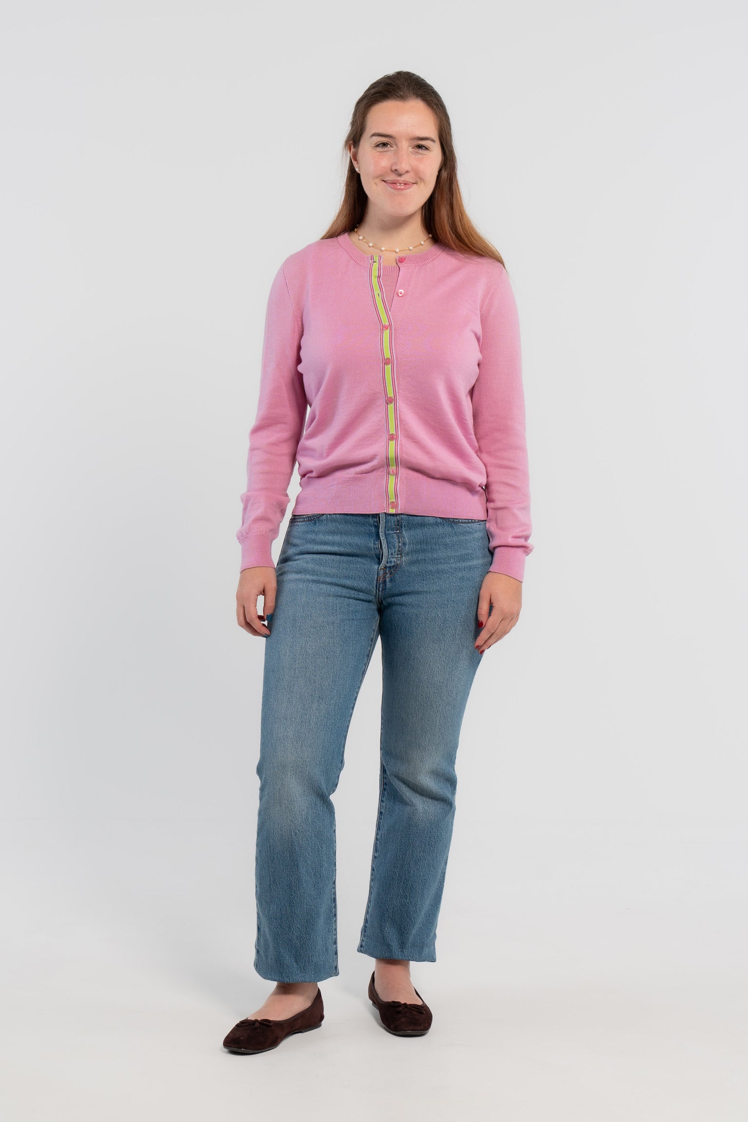 Woman wearing orchid pink cardigan with pink buttons and green, pink, lilac striped grosgrain ribbon trim, pearl choker necklace, jeans, ballet flats on plain background.