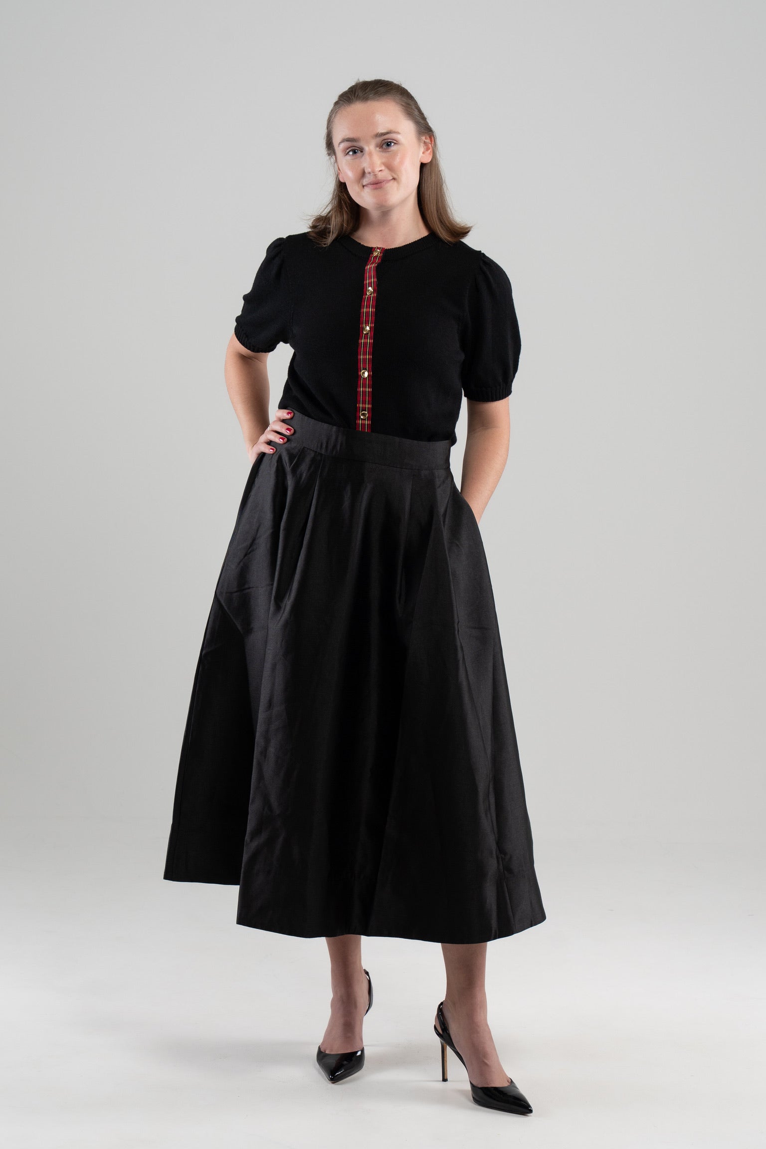 Woman wearing black puff-sleeve cardigan with red tartan plain ribbon trim and gold buttons, black skirt and black patent sling-back shoes on plain background.